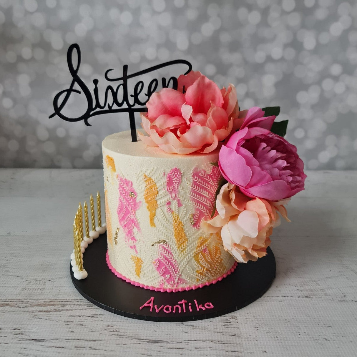 Decorative cake with floral elements and 'Sixteen' topper on a textured surface with blurred background
